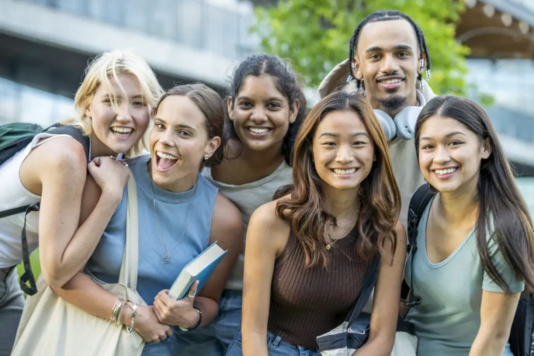 A small group of six University students huddle in closely together as they pose for a portrait. They are each dressed casually and have backpacks on and textbooks in hand as they smile while spending time outside n campus.