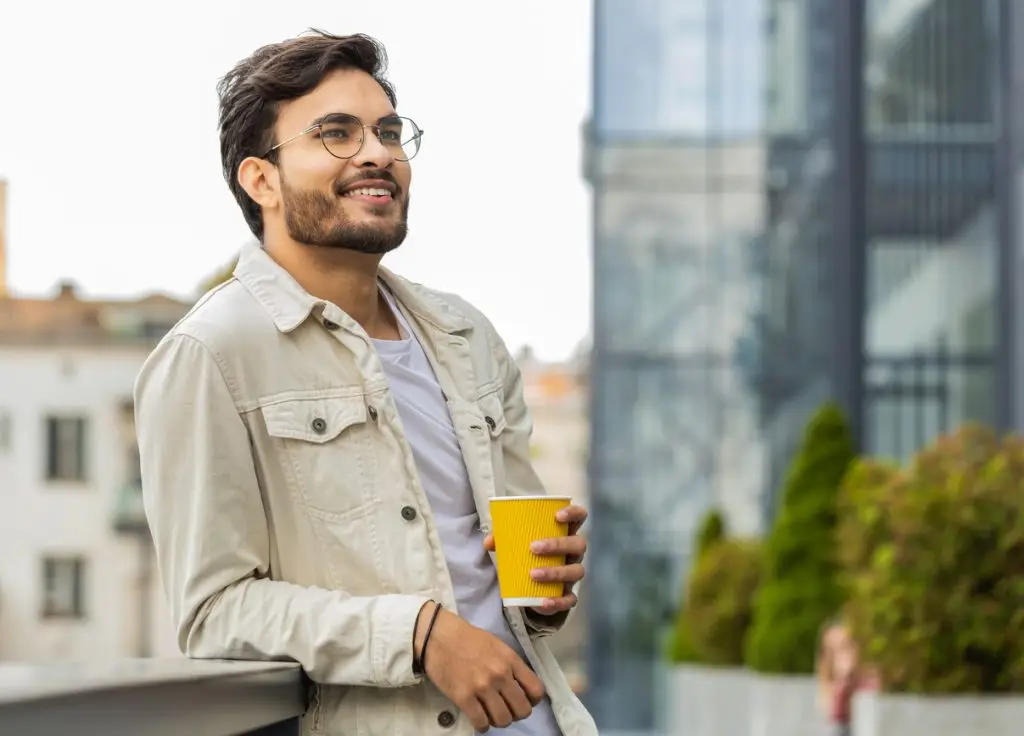 Happy Indian man enjoying drinking morning coffee hot drink, relaxing, taking a break in city street